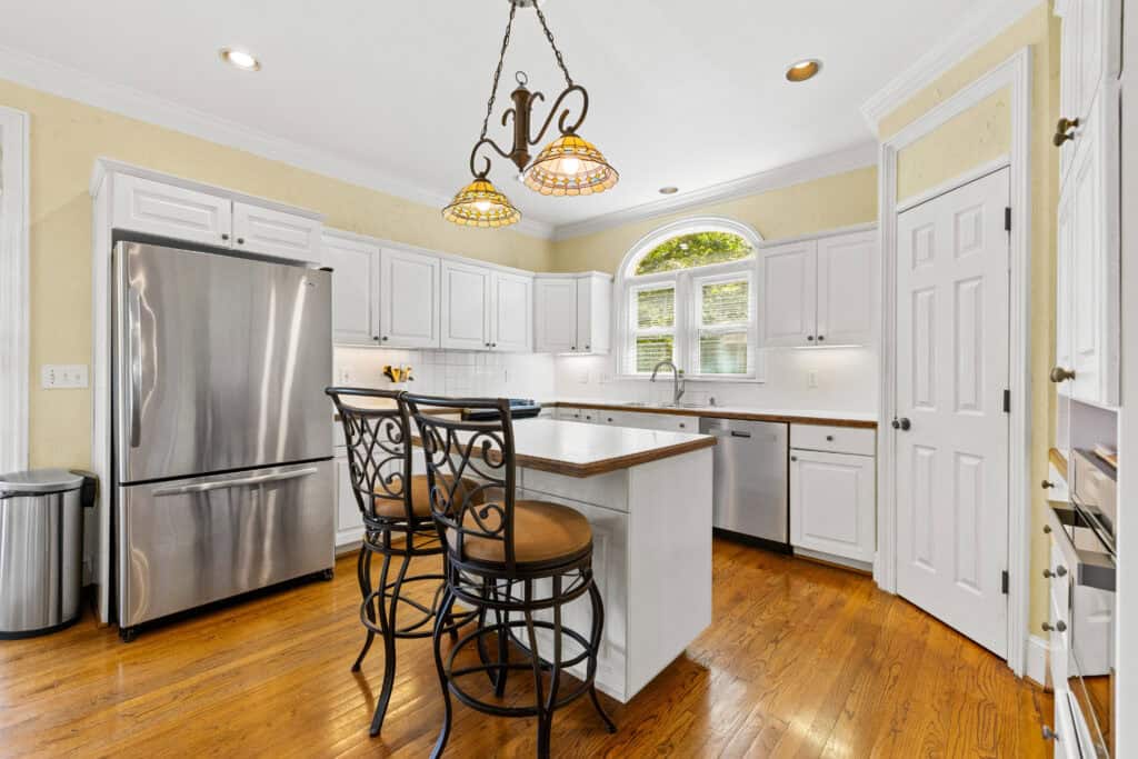 Bright white kitchen with stainless steel refrigerator, central island, pendant lights, and hardwood flooring.