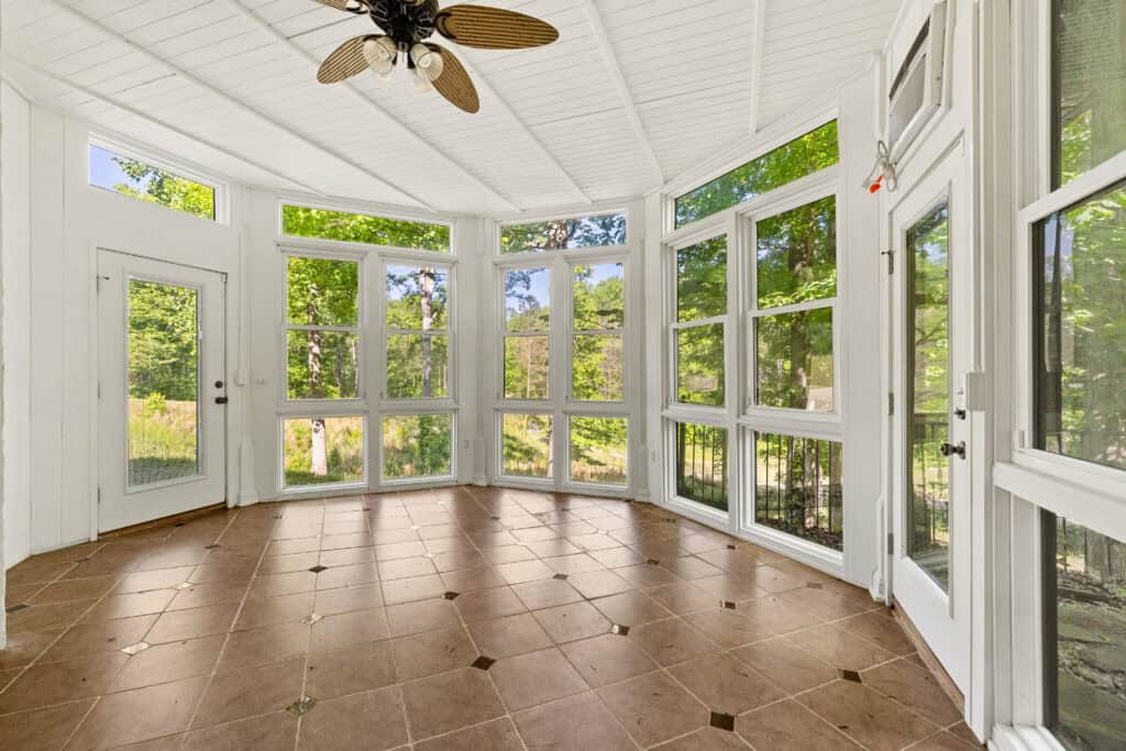 Bright enclosed sunroom with tiled floor, ceiling fan, and wraparound windows overlooking trees.