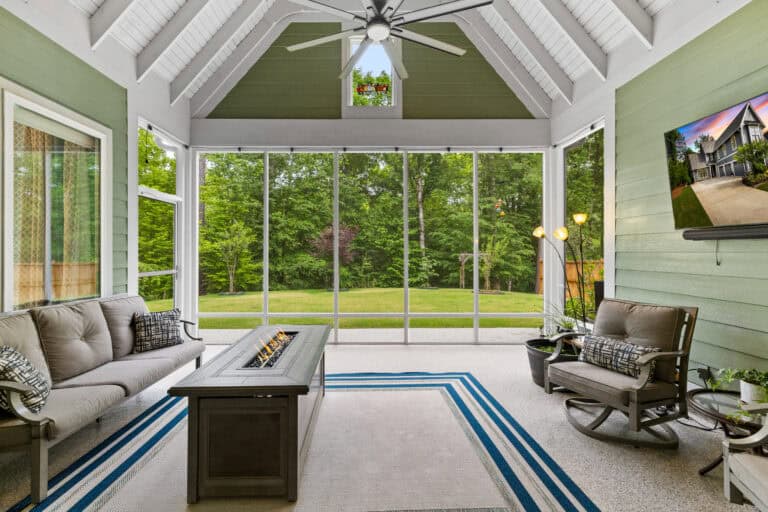 Bright enclosed sunroom with vaulted ceiling, seating area, ceiling fan, and views of a wooded backyard.