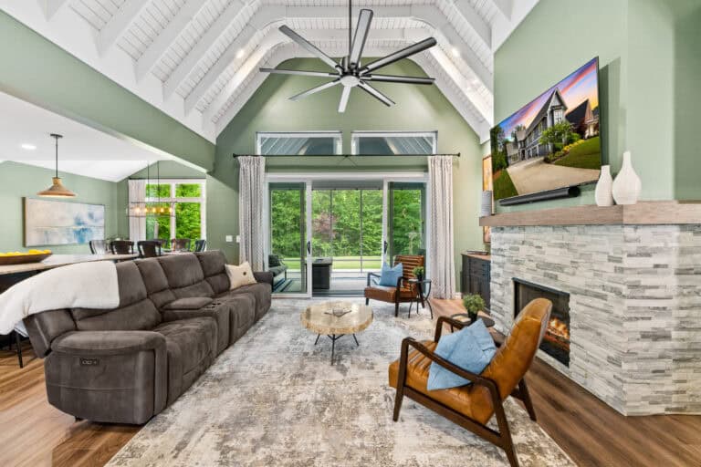 Bright living room with vaulted ceiling, fireplace, furnishings, and wooded views through glass doors.