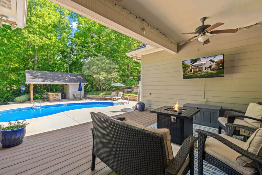 Covered outdoor patio with seating, fire table, and swimming pool in a wooded backyard
